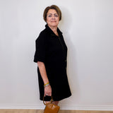 Woman in a black coat holding a brown handbag against a white wall.