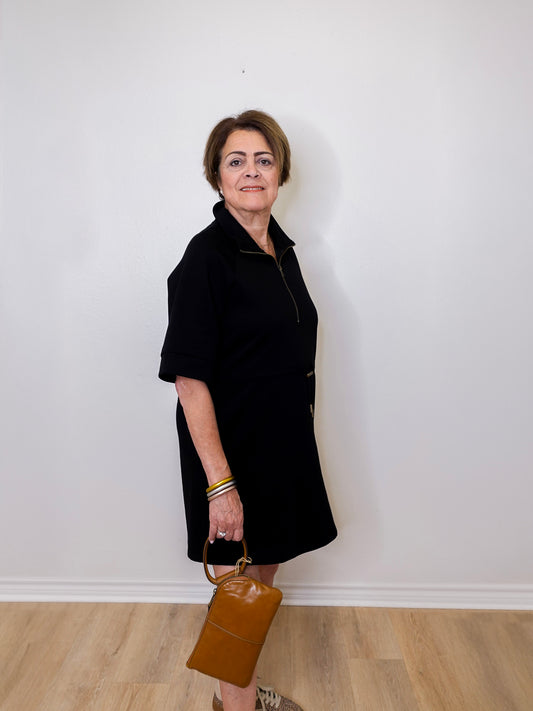 Woman in a black coat holding a brown handbag against a white wall.