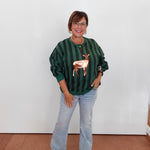 A woman wearing a dark green and black vertically striped long-sleeve top with a metallic rose gold deer applique on the front, paired with light-wash blue jeans and beige sandals, standing against a plain white wall.