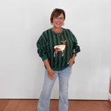 A woman wearing a dark green and black vertically striped long-sleeve top with a metallic rose gold deer applique on the front, paired with light-wash blue jeans and beige sandals, standing against a plain white wall.