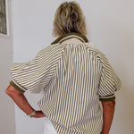 Person wearing a striped shirt and white pants standing against a plain wall.