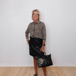 Person wearing a leopard print shirt and black leather skirt, holding a black handbag against a white wall.