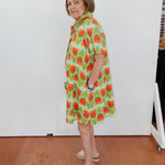 Woman wearing a colorful dress with a pattern of watermelons in a room with a white wall and tiled floor.