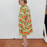 Woman wearing a colorful dress with a pattern of watermelons in a room with a white wall and tiled floor.