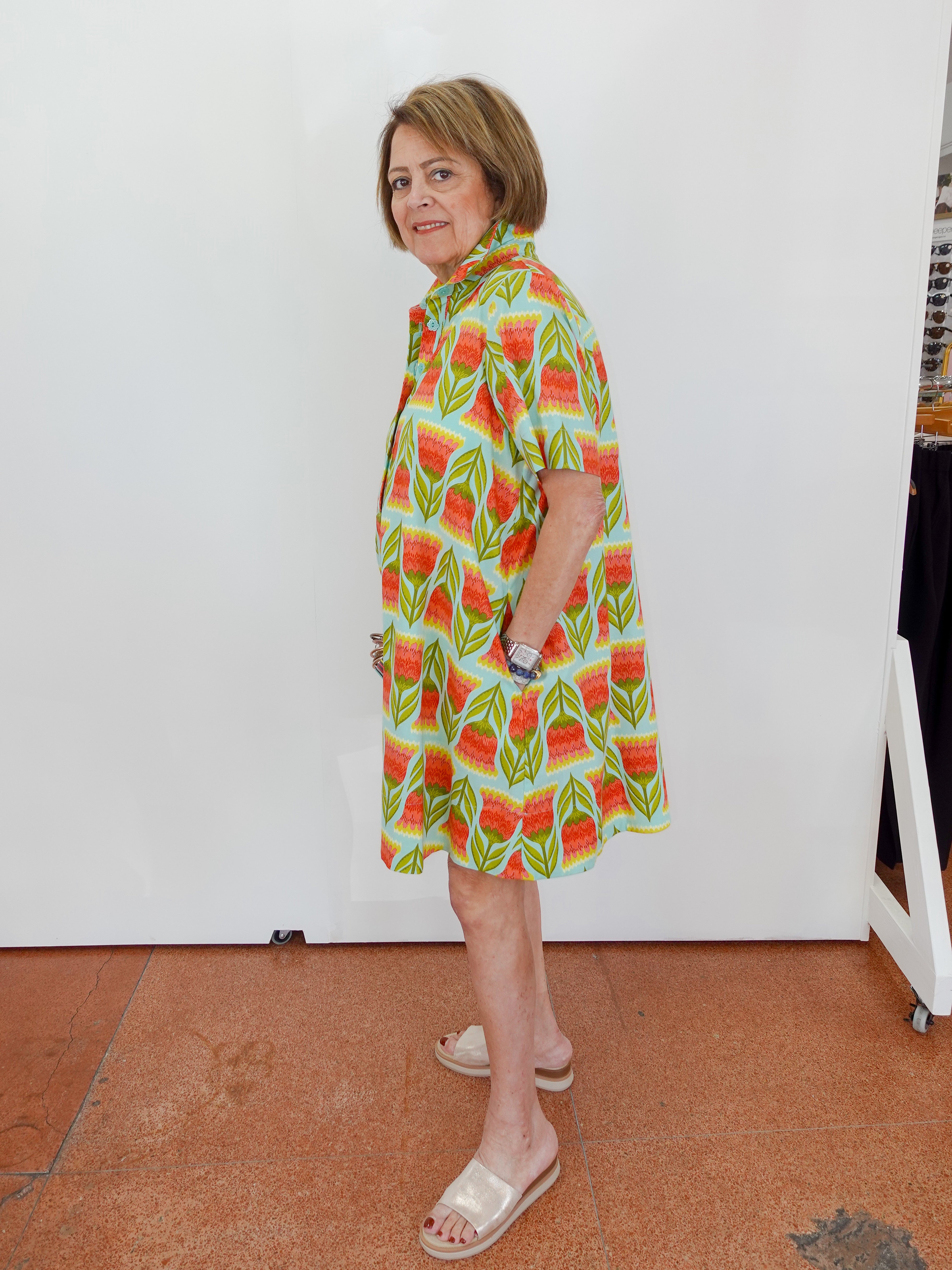 Woman wearing a colorful dress with a pattern of watermelons in a room with a white wall and tiled floor.