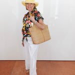 Woman wearing a floral shirt, white pants, yellow hat, and holding a straw bag on a tiled floor.