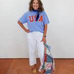 Woman wearing a blue 'USA' t-shirt and white pants, holding a colorful patterned bag against a white wall.