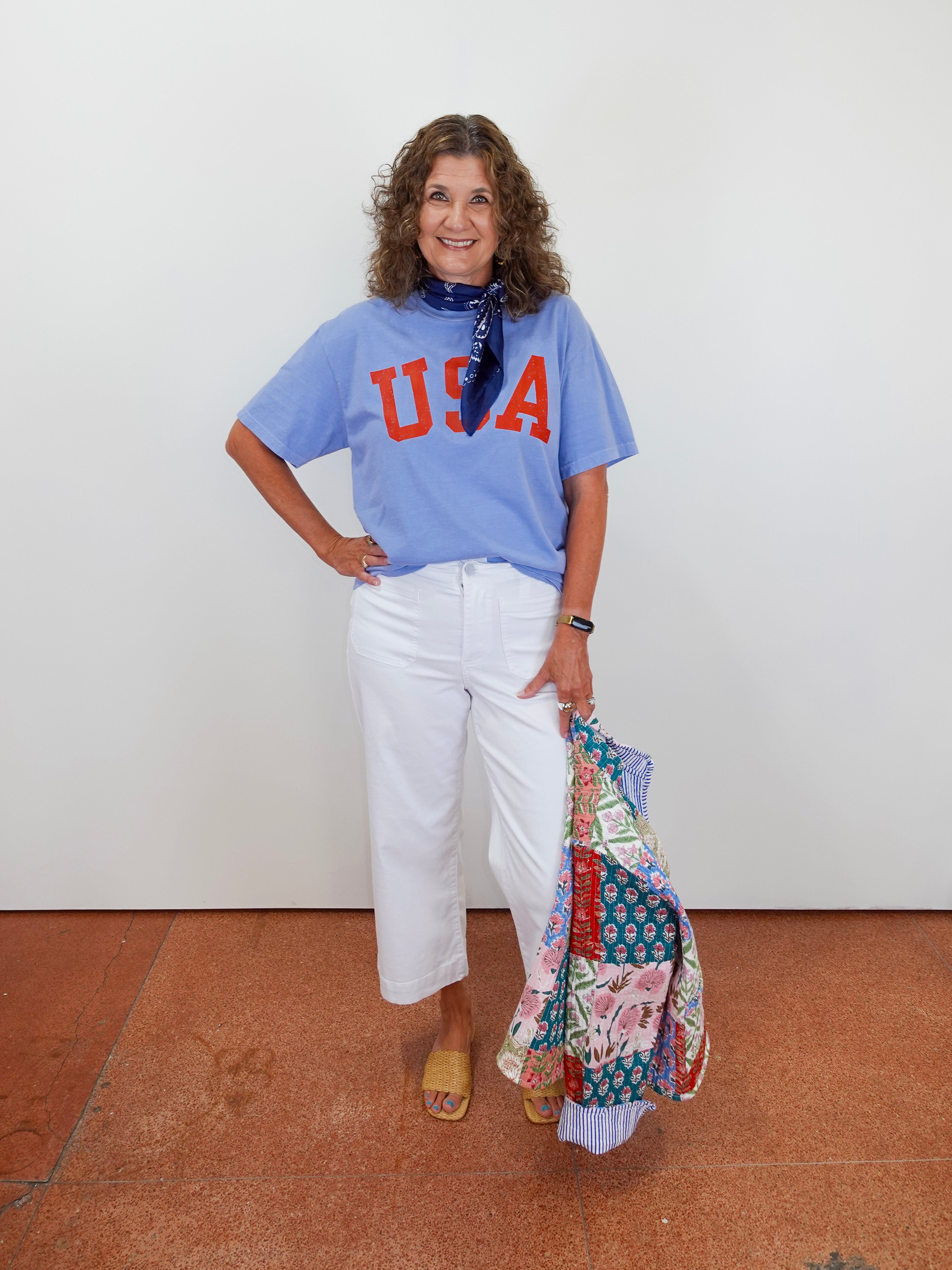 Woman wearing a blue 'USA' t-shirt and white pants, holding a colorful patterned bag against a white wall.