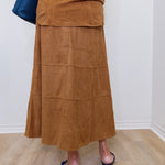 Person wearing a brown suede skirt and navy shoes on a light wooden floor.