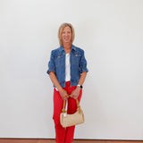 Woman wearing a denim jacket, white shirt, red pants, and holding a beige handbag against a white background.