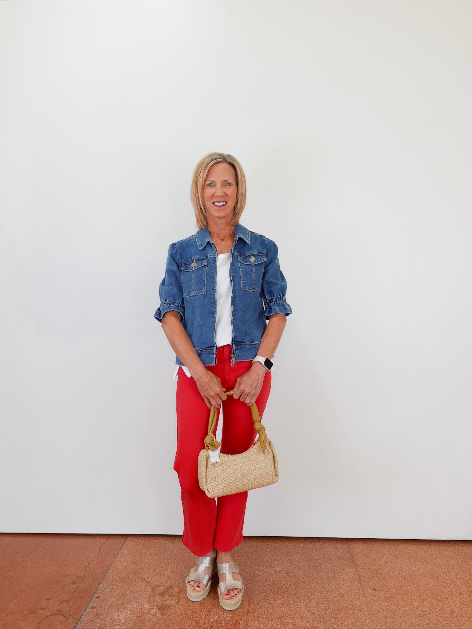 Woman wearing a denim jacket, white shirt, red pants, and holding a beige handbag against a white background.