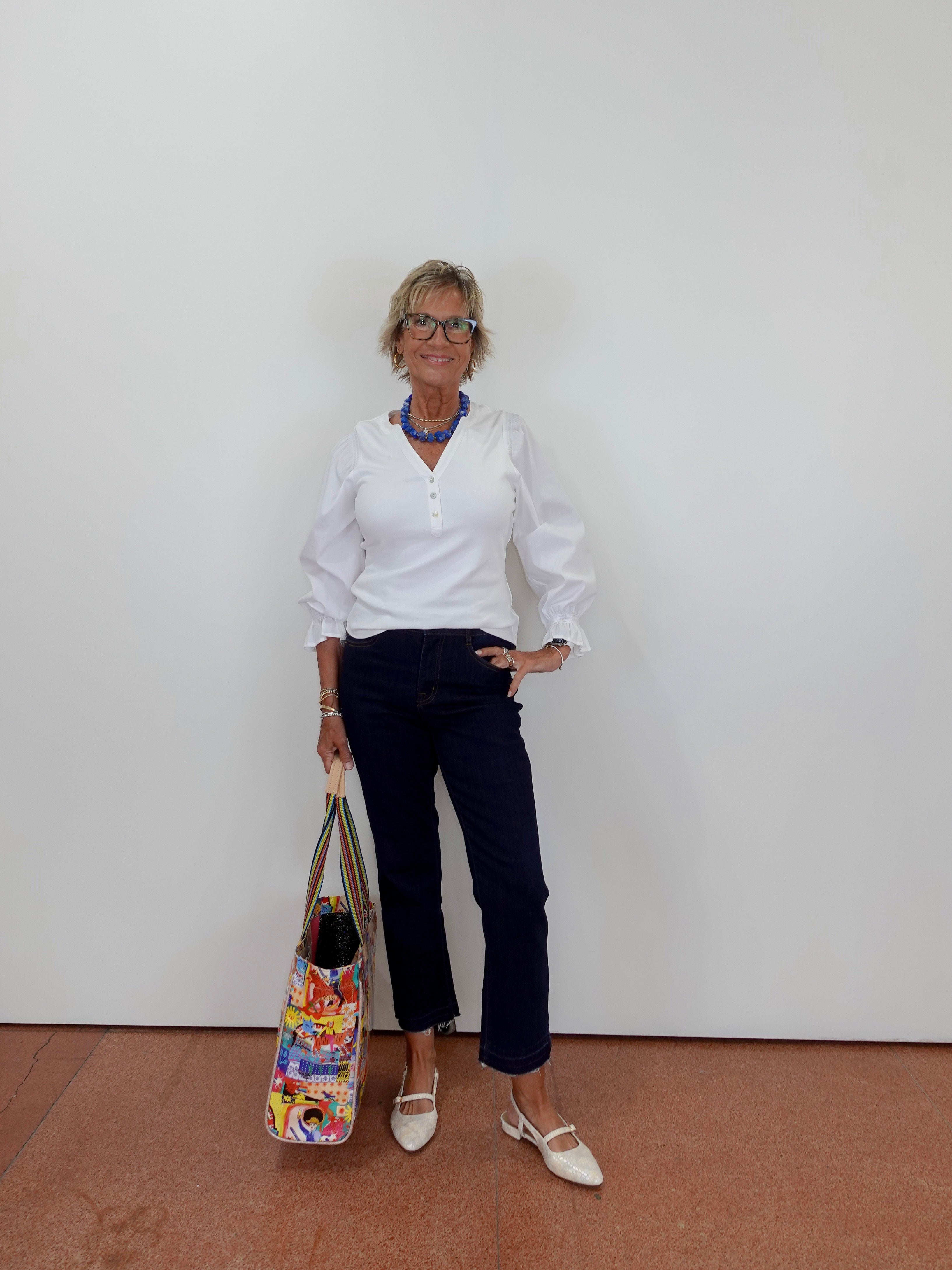 Woman in white shirt and navy pants holding a colorful bag against a plain background