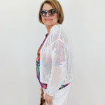 Woman wearing a white crochet cardigan over a colorful top with sunglasses and a handbag on a plain background