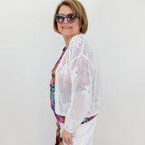 Woman wearing a white crochet cardigan over a colorful top with sunglasses and a handbag on a plain background