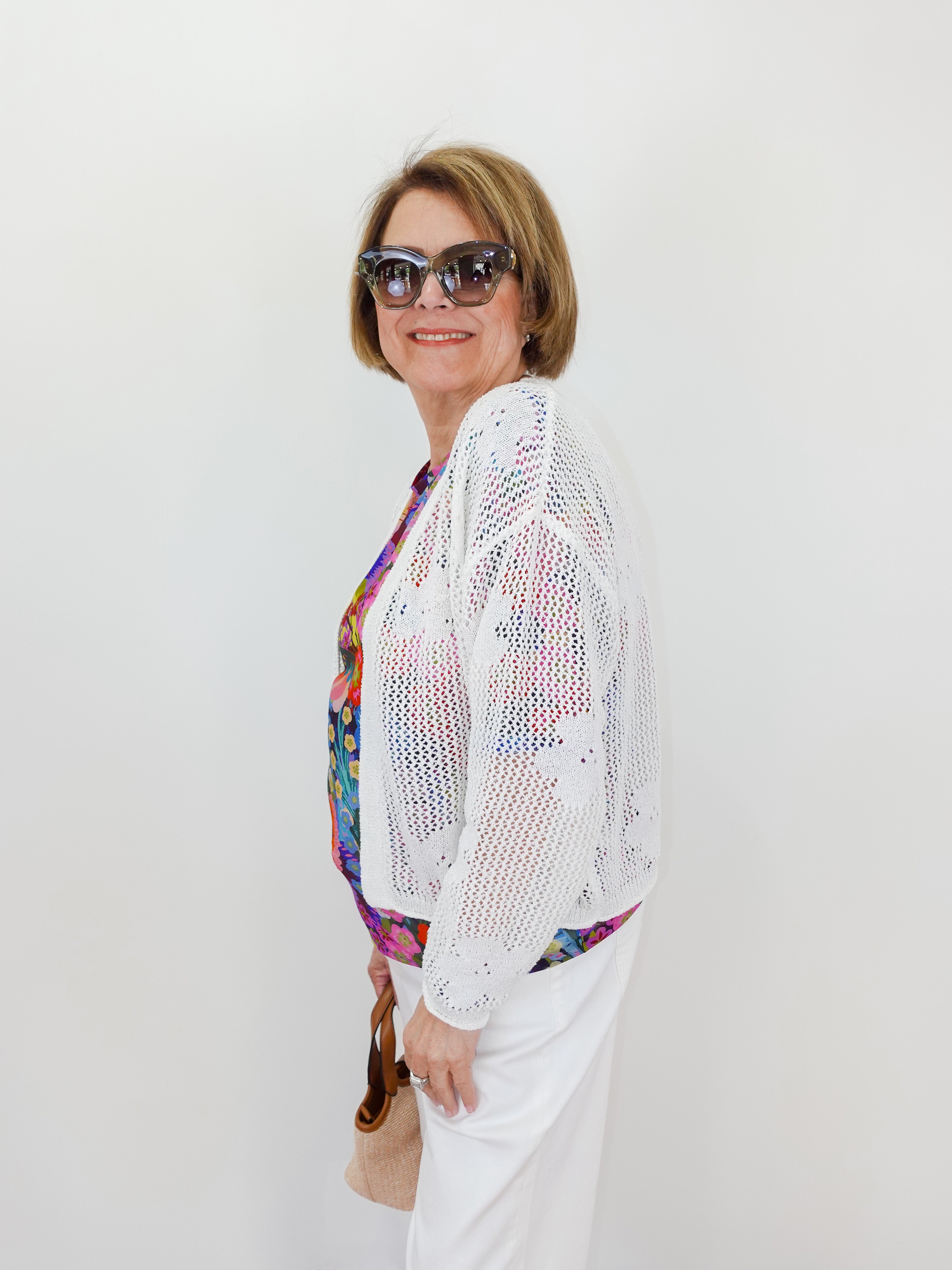 Woman wearing a white crochet cardigan over a colorful top with sunglasses and a handbag on a plain background