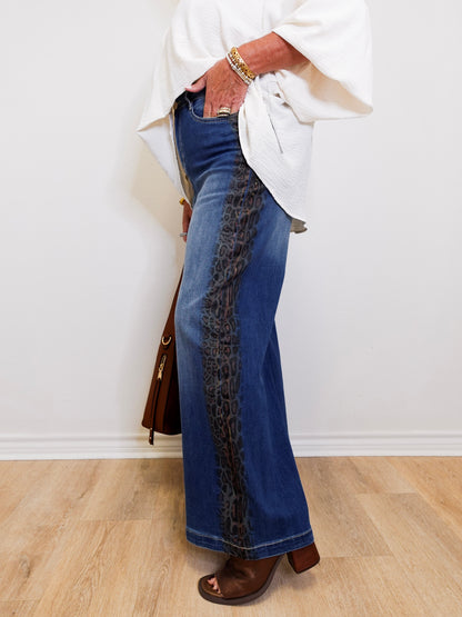 Person wearing blue jeans with a leopard print side panel, standing against a white wall.