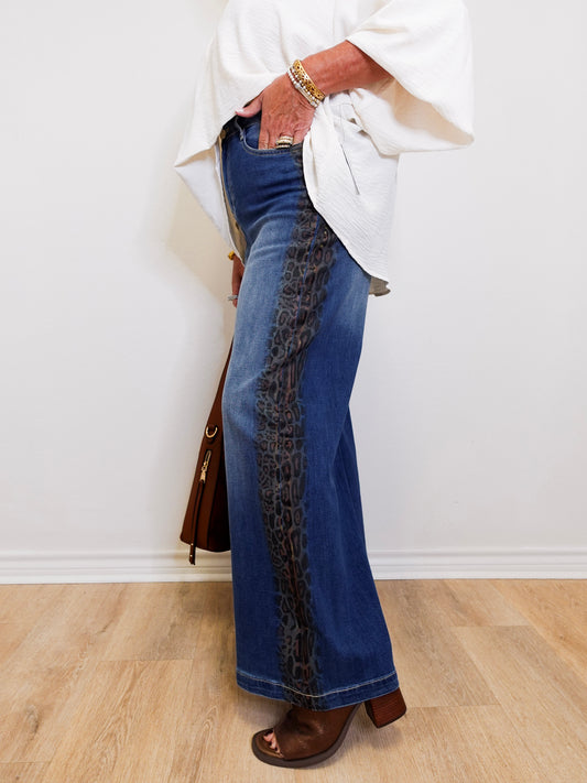 Person wearing blue jeans with a leopard print side panel, standing against a white wall.