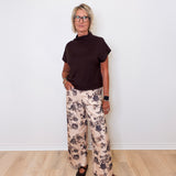 Person wearing a black top and floral pants standing against a white wall.