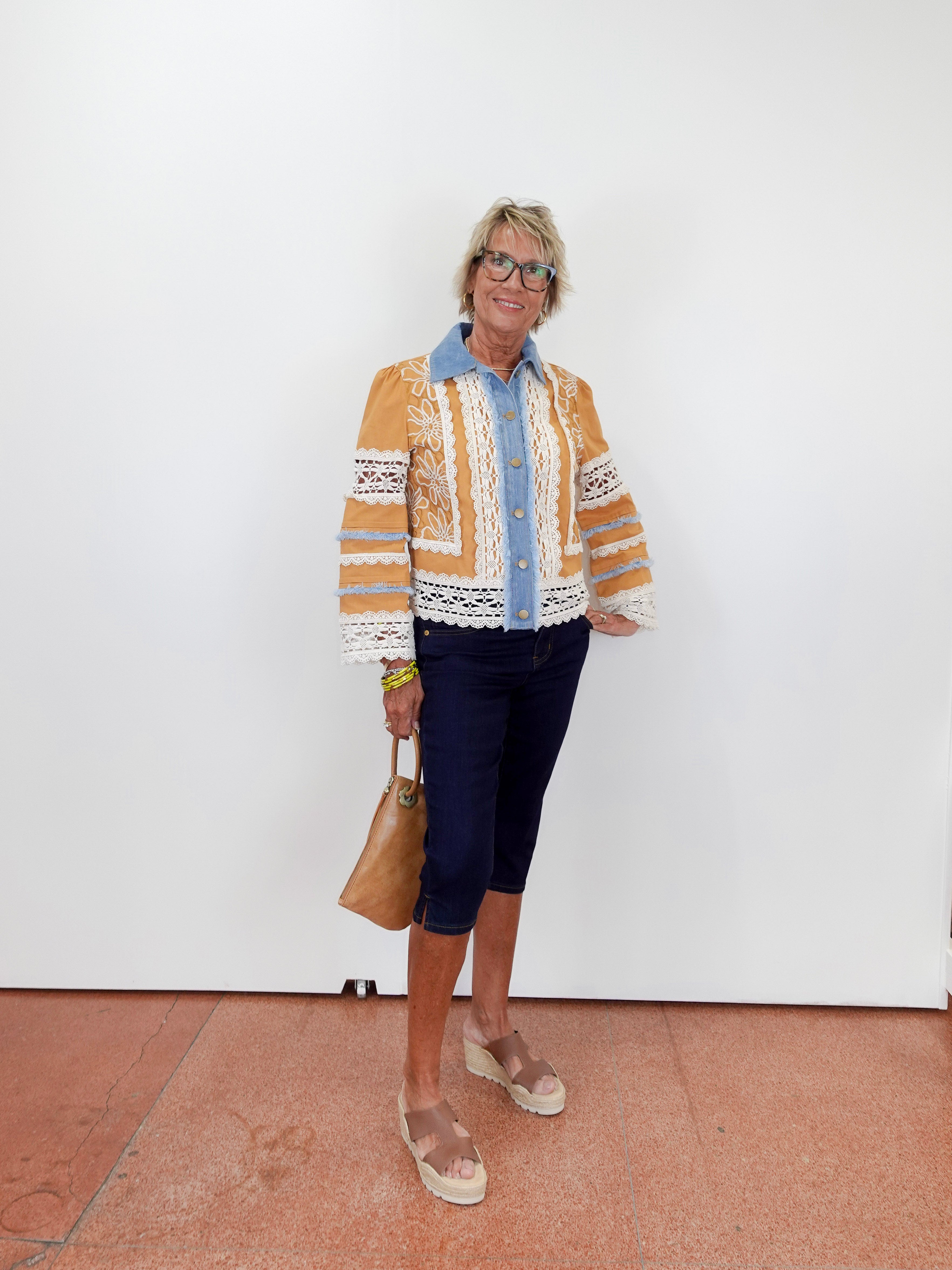 Woman wearing a patterned cardigan over a shirt with dark pants, standing against a white wall.