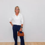Woman wearing a white sweater and blue jeans, holding a brown handbag against a plain background