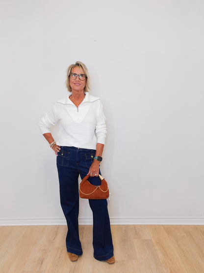 Woman wearing a white sweater and blue jeans, holding a brown handbag against a plain background