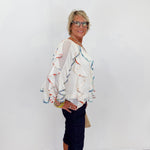 Woman wearing a colorful blouse and dark pants against a white wall.