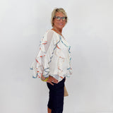 Woman wearing a colorful blouse and dark pants against a white wall.