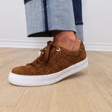 Brown suede sneaker with white sole worn by a person on a wooden floor.