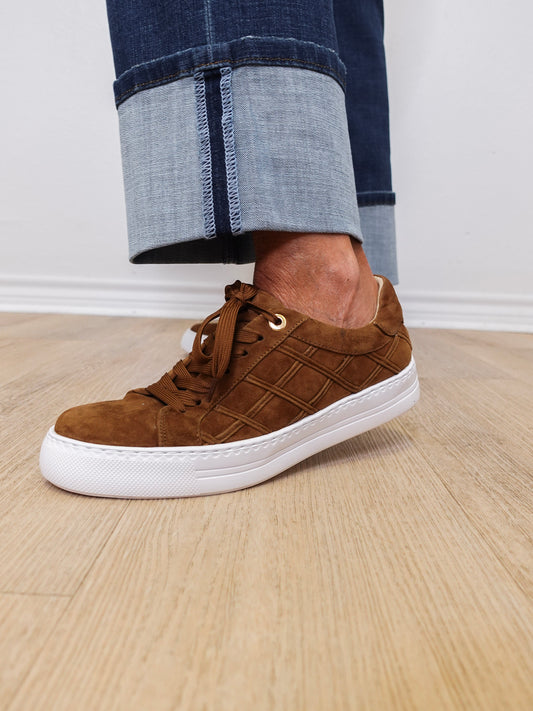 Brown suede sneaker with white sole worn by a person on a wooden floor.