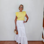 Woman wearing a yellow top and white skirt against a white wall.