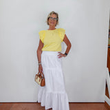 Woman wearing a yellow top and white skirt against a white wall.