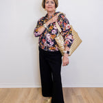 Woman wearing a floral blouse and black pants, holding a gold handbag against a white wall.
