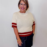 Woman wearing a cream sweater with rust accents and a denim skirt against a white wall.