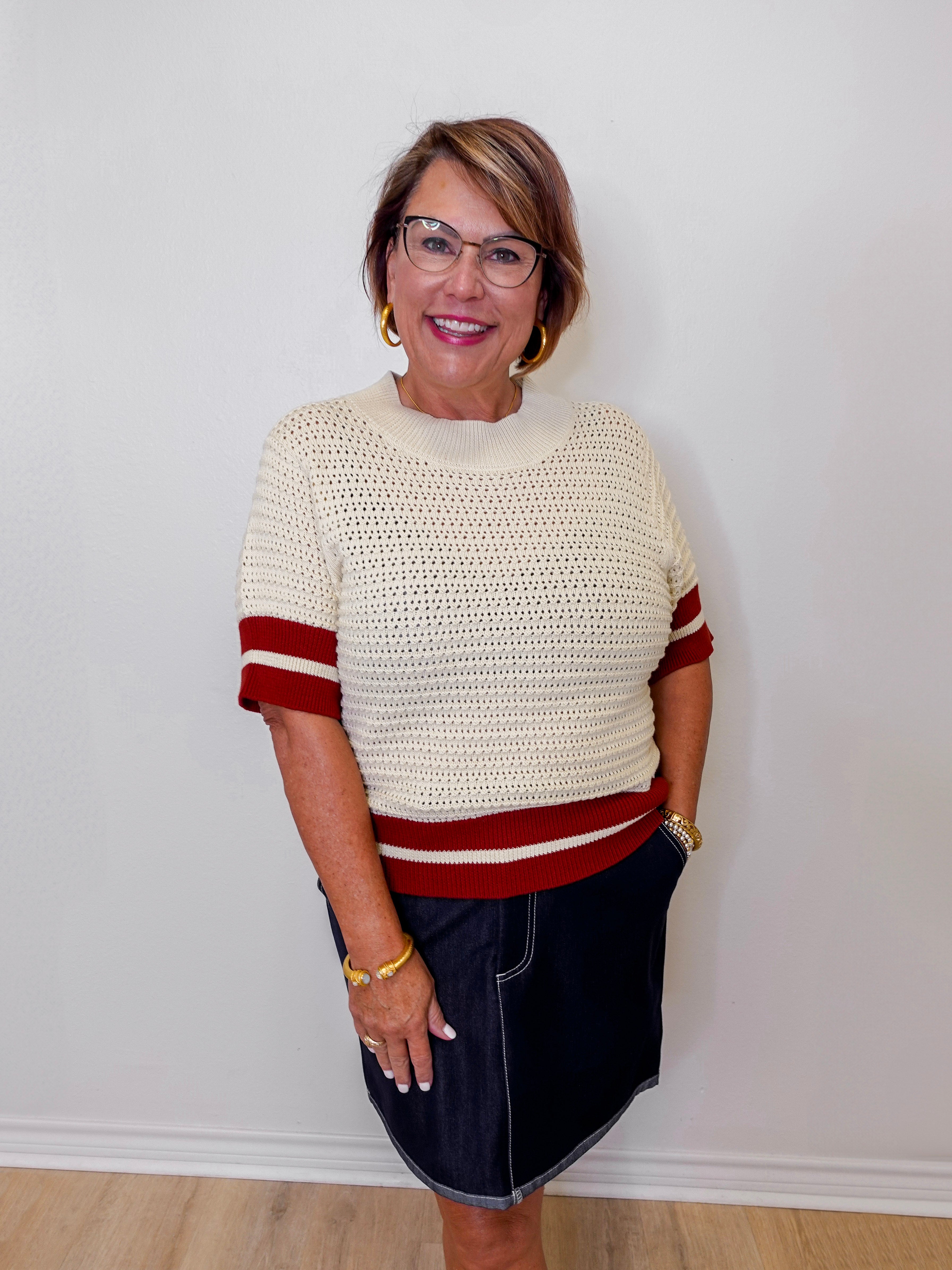 Woman wearing a cream sweater with rust accents and a denim skirt against a white wall.