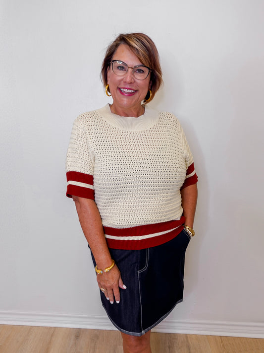 Woman wearing a cream sweater with rust accents and a denim skirt against a white wall.